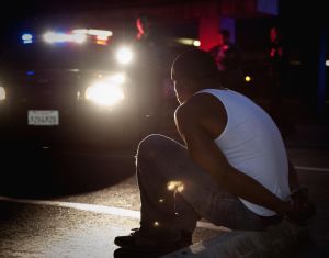 Police shining lights on handcuffed African man sitting on curb