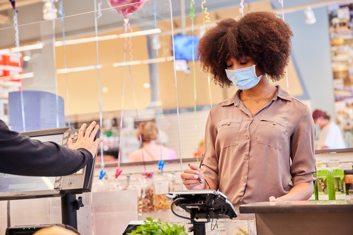 Woman in a face mask paying for her groceries at a plexiglass protected checkout