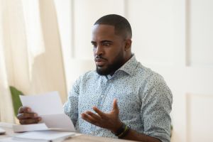 African man sitting at table holding letter reading awful news