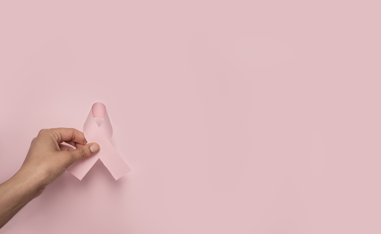 Cropped hand holding a pink awareness ribbon against a pink background, symbolizing breast cancer awareness and support