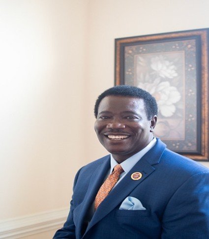 A smiling African American man in a blue suit and orange tie standing in front of a framed artwork.