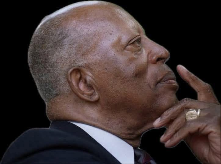 A close-up portrait of a bald, middle-aged Black man in a suit, looking thoughtful with his hand on his chin.