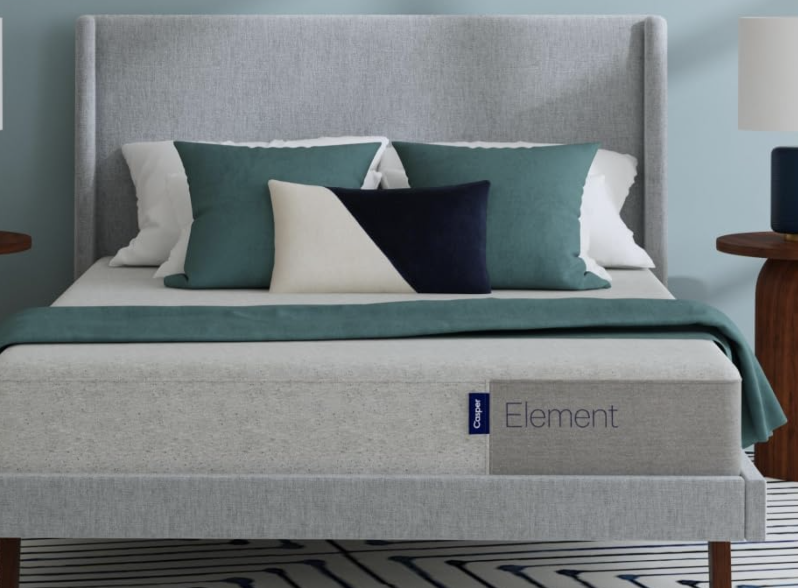 A gray upholstered bed with green, white, and black throw pillows. The bed has a gray headboard and is positioned in front of a light blue wall. A gray box labeled "Element" is visible at the foot of the bed.