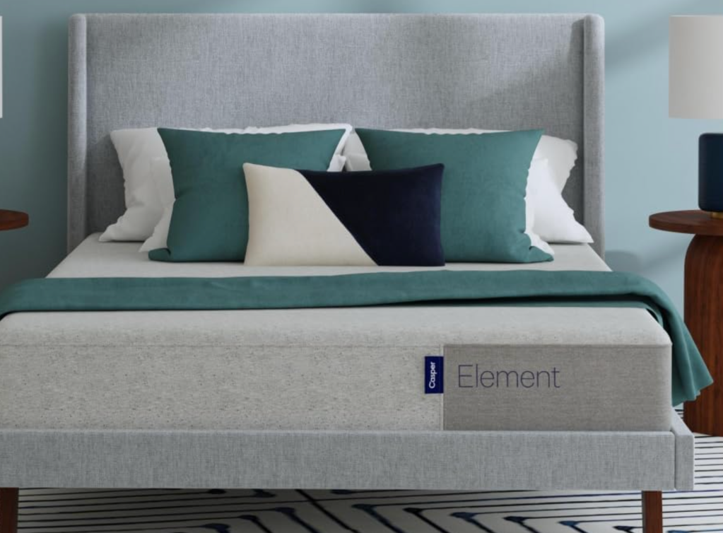 A gray upholstered bed with green, white, and black throw pillows. The bed has a gray headboard and is positioned in front of a light blue wall. A gray box labeled "Element" is visible at the foot of the bed.