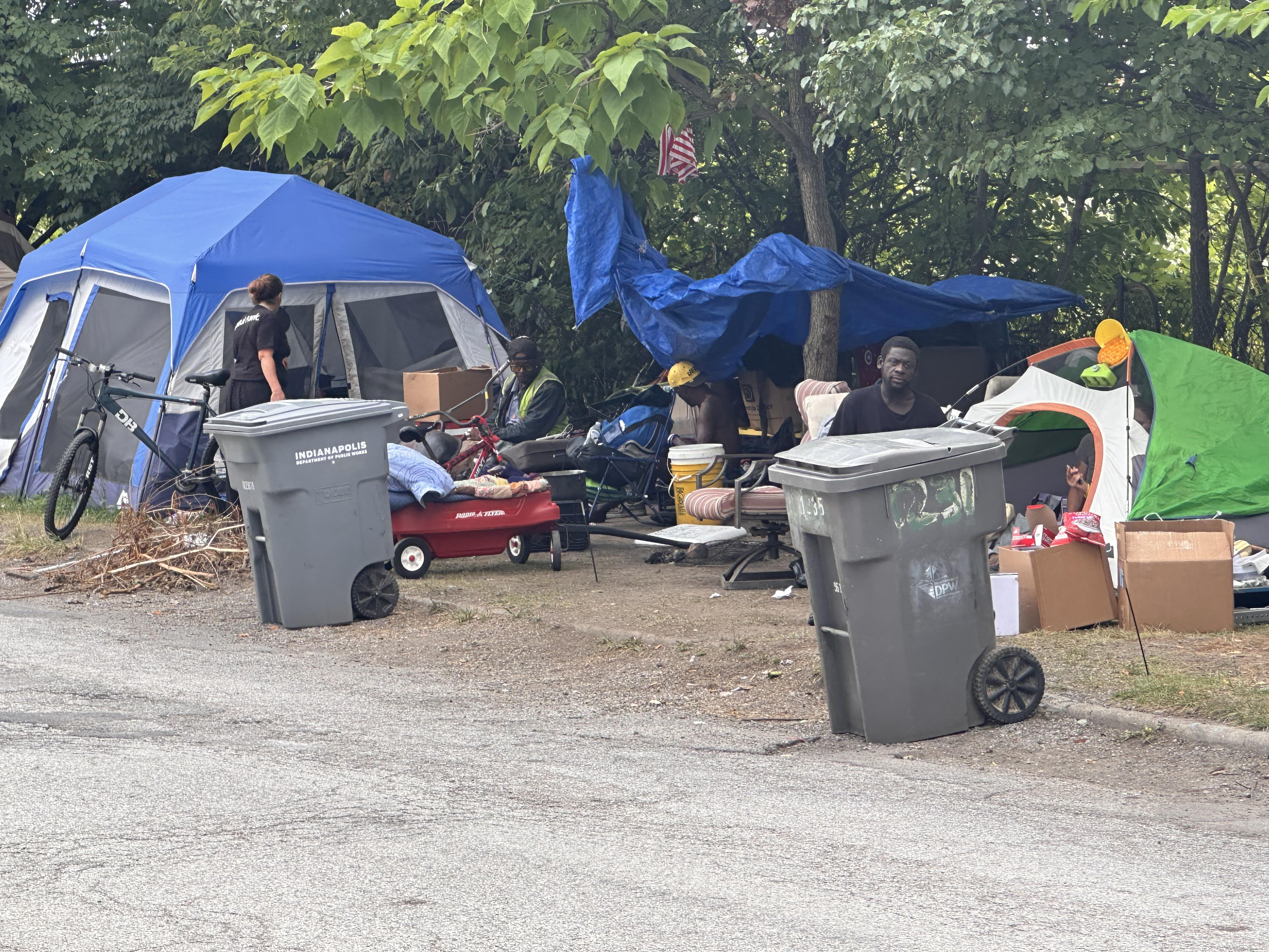 Outdoor campsite with tents, trash cans, and personal belongings scattered on the ground. An American flag is visible in the trees.