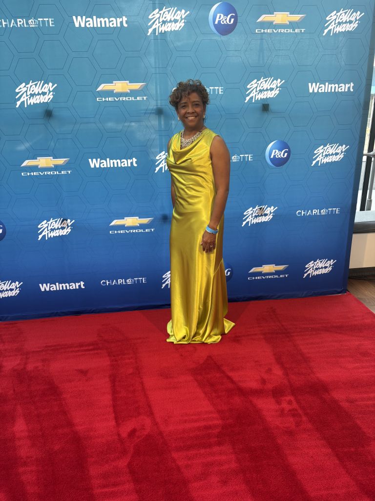 A smiling woman wearing a yellow dress stands on a red carpet in front of a backdrop featuring the Stellar Awards, Walmart, Chevrolet, and P&G logos.