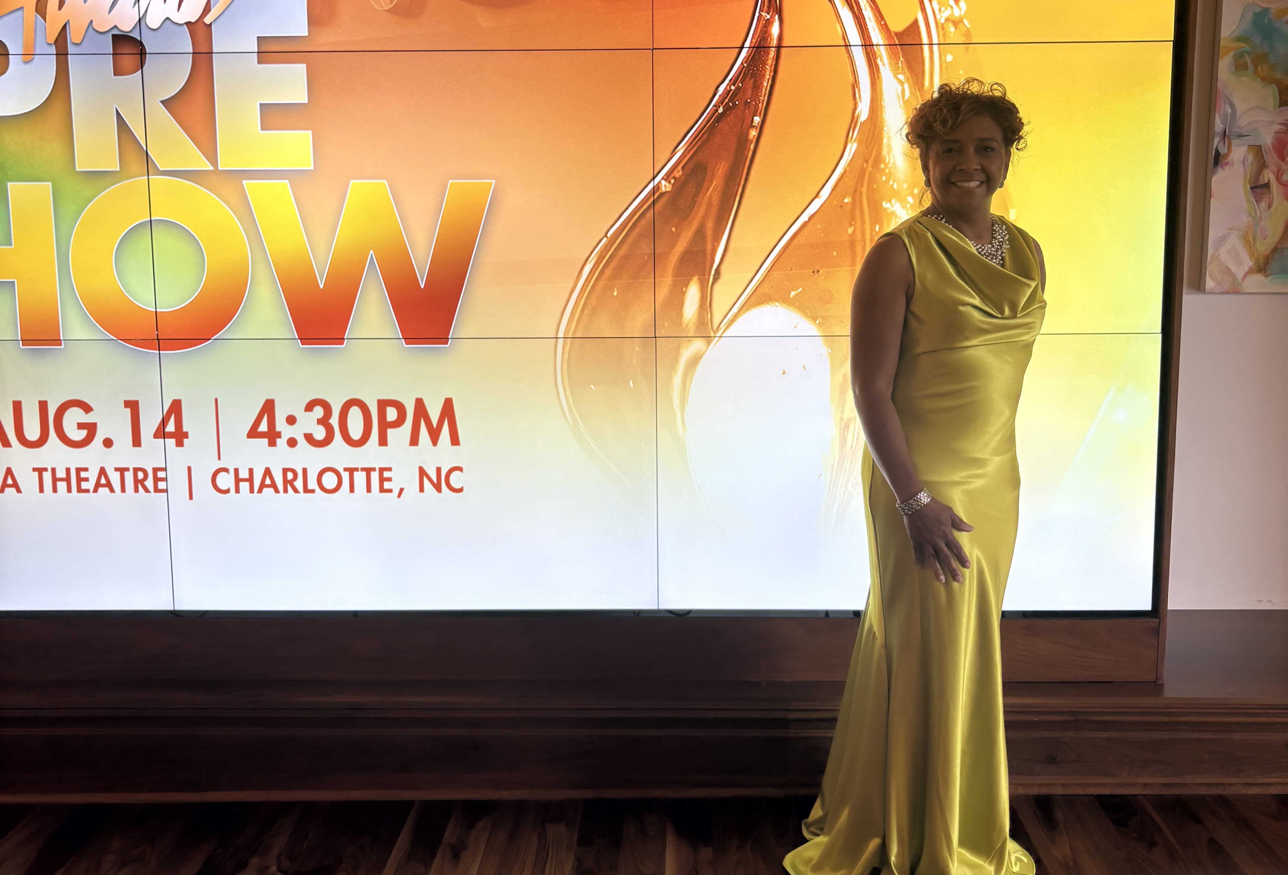 A smiling woman in a yellow dress stands in front of a large display advertising the "Annual Stellar Awards" event on August 14 at 4:30 PM in Charlotte, NC.