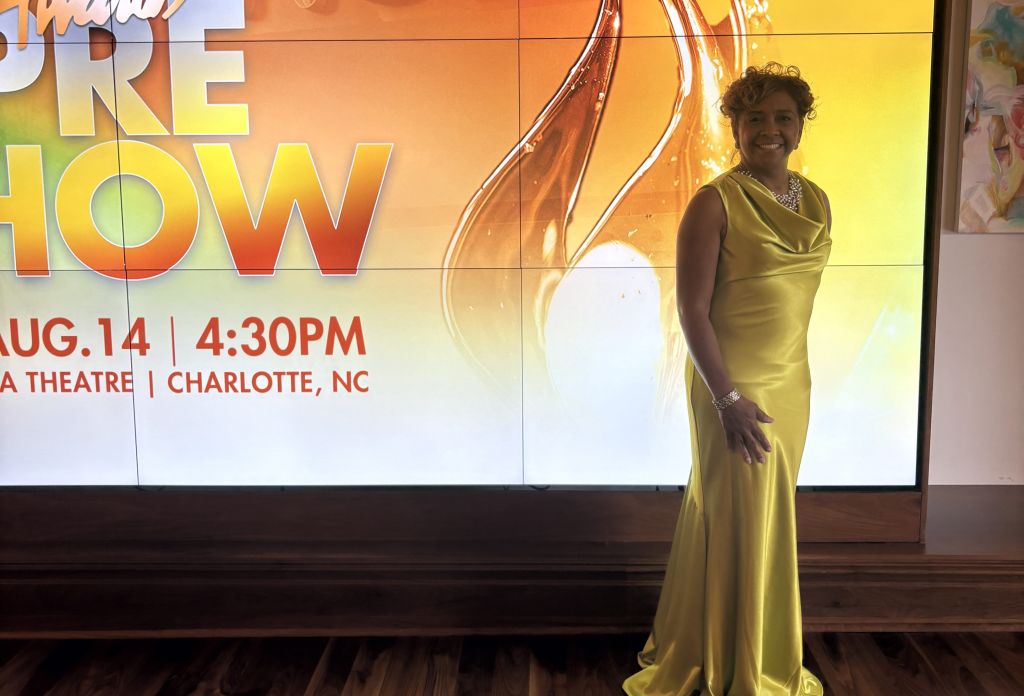 A smiling woman in a yellow dress stands in front of a large display advertising the "Annual Stellar Awards" event on August 14 at 4:30 PM in Charlotte, NC.