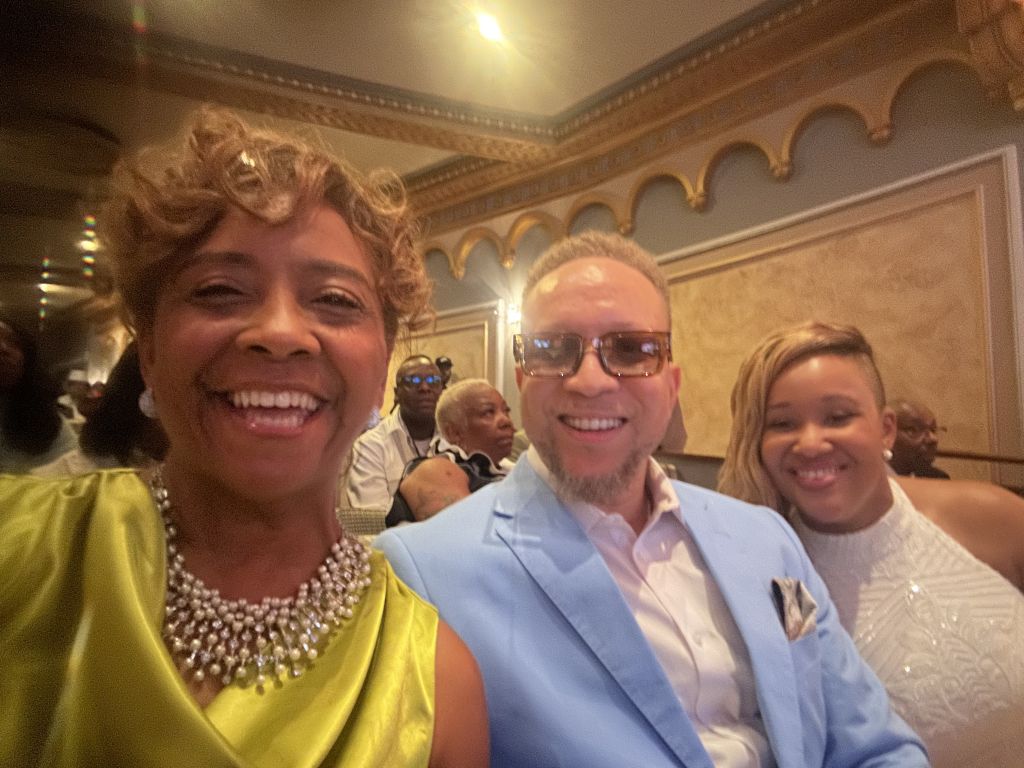 Three smiling people posing together in an ornate, gilded room.