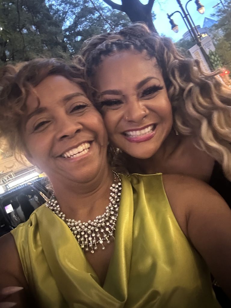 Two smiling women with curly hair wearing colorful dresses and jewelry, standing outdoors in front of trees.