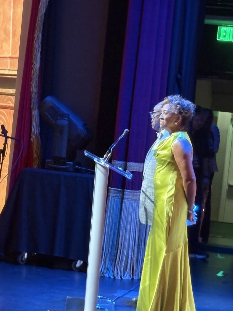 A woman in a yellow dress standing at a podium on a stage with blue curtains and an exit sign visible.