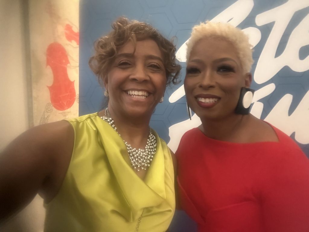 Two smiling women, one with curly brown hair and the other with short blonde hair, posing together in front of a colorful mural.